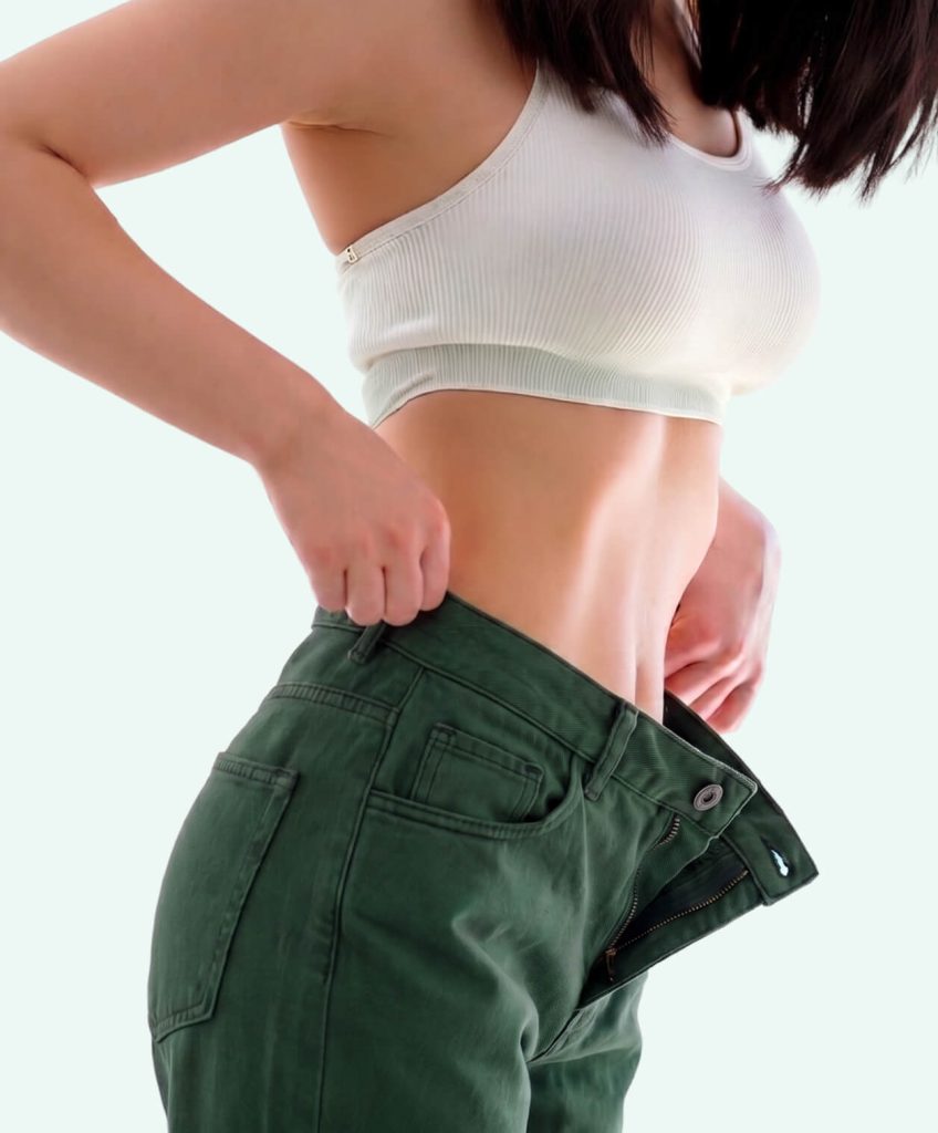 Woman pulling away the loose waistband of her green jeans, demonstrating a flat stomach and no bloating after passing the zipper test.